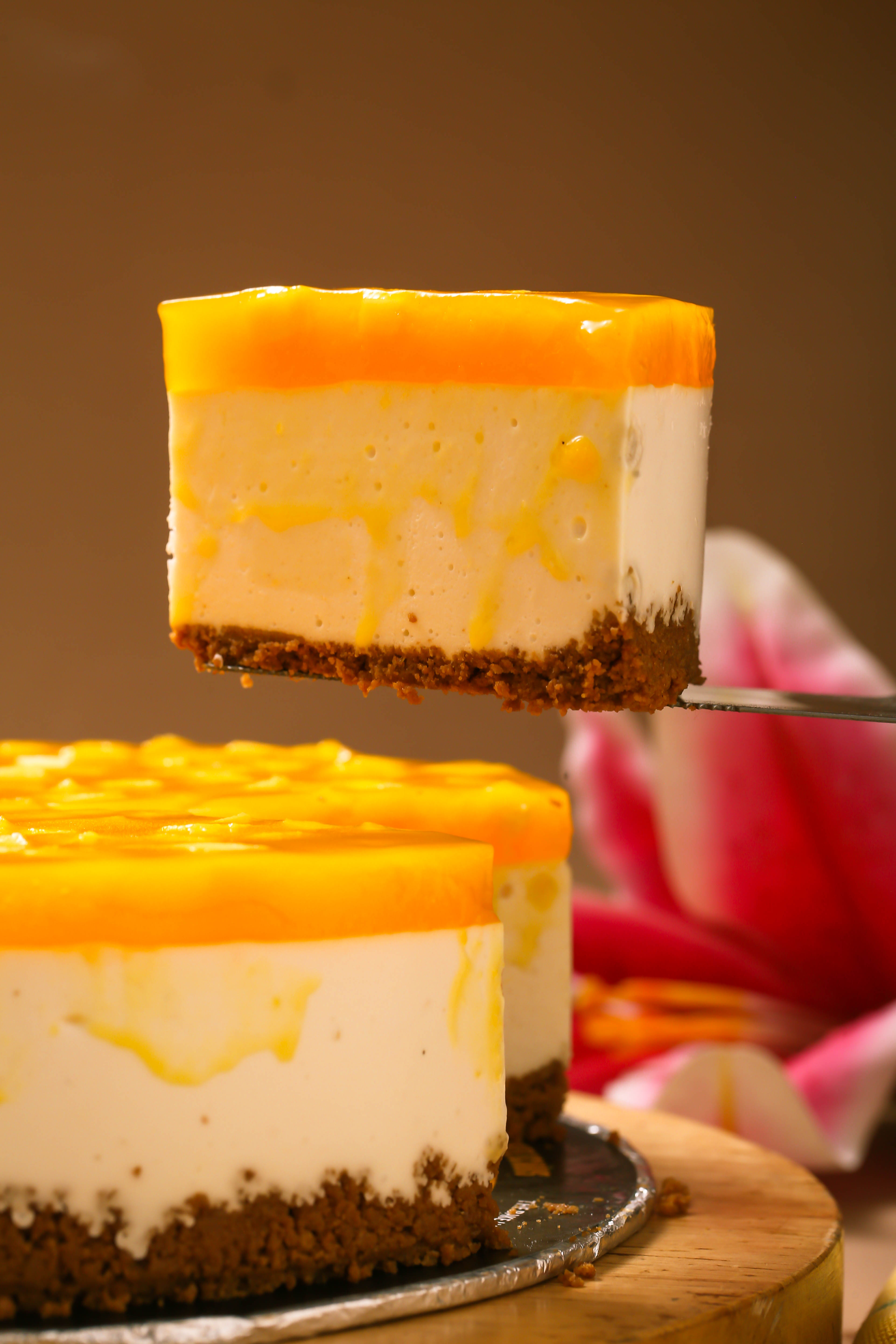 Creamy mango cheesecake slice with glossy topping and biscuit crust on a serving spatula